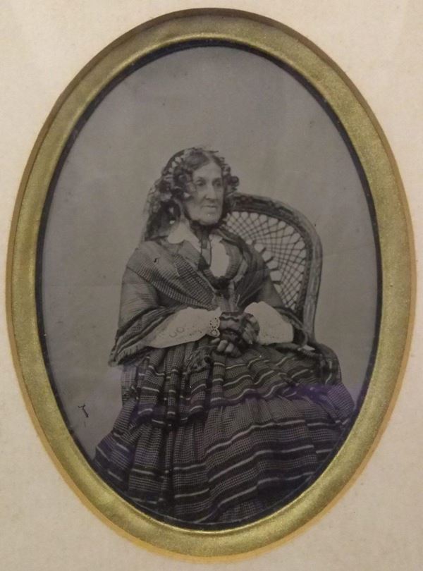 Photo of Elizabeth Glasson in an oval frame. She is sitting very upright in a wicker chair, wearing heavy Victorian clothes: a layered dress with horizontal stripes a white broderie anglaise blouse and black lace gloves and headdress. Her hair is curled and styled into ringlets around her face.,